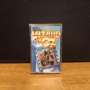 May include: A vintage cassette tape of "The Wizard of Oz" original cast album. The cover features the film's title in gold lettering and a scene from the movie, starring Judy Garland. The cassette is in a clear plastic case.