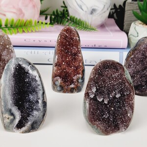 May include: Five amethyst crystal geodes in varying shades of purple, brown, and gray. The geodes have a rough, textured exterior and a sparkling, crystalline interior. The crystals are displayed on a white surface.