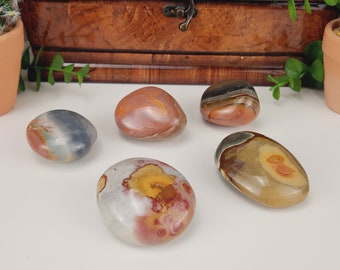 Puede incluir: Cinco piedras pulidas en tonos marrón, rojo, gris y amarillo. Las piedras tienen formas lisas y redondeadas y patrones variados. El fondo incluye una caja de madera, pequeñas plantas en macetas y una superficie blanca.