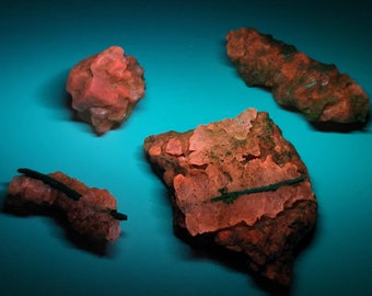 Może przedstawiać: Cztery kawałki surowych, grubo ciętych minerałów o świecącej pomarańczowej barwie, prawdopodobnie fluorescencyjne. Minerały mają teksturowaną powierzchnię i są umieszczone na turkusowym tle. Niektóre kawałki mają cienkie, ciemnozielone linie.