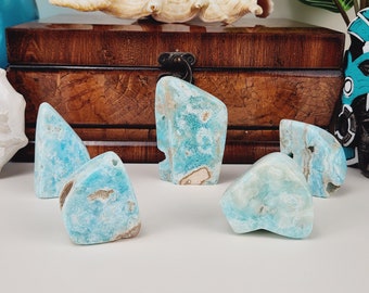 Könnte beinhalten: Eine Auswahl an Larimar-Edelsteinen, die eine Mischung aus hellblauen und weißen Farben aufweisen, mit natürlichen, unregelmäßigen Formen. Einige Steine haben braune Markierungen. Im Hintergrund befinden sich eine Holzkiste und andere dekorative Elemente.