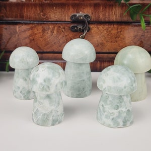 May include: Five light green mushroom figurines with a speckled pattern. The mushroom caps are rounded, and the stems are cylindrical. The mushrooms are arranged on a white surface, with a wooden box in the background.