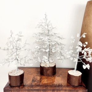 Könnte beinhalten: Drei dekorative Kristallbäume mit silbernen Drahtzweigen und klaren Kristallblättern. Die Bäume stehen in kleinen, braunen, zylindrischen Töpfen, die mit kleinen Steinen gefüllt sind. Die Bäume stehen auf einer dunklen Holzoberfläche.
