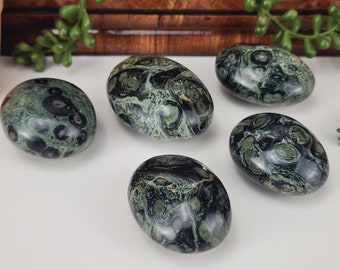 May include: Five polished oval stones with a dark green and black mottled pattern. The stones have a smooth, glossy finish and are arranged on a white surface. The stones are a natural product.
