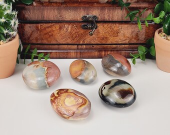 Puede incluir: Cinco piedras pulidas en tonos marrón, beige y gris están dispuestas sobre una superficie blanca. Las piedras tienen formas lisas y redondeadas y patrones variados. Una caja de madera y plantas en macetas están en el fondo.