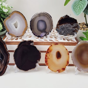 May include: Seven agate slices in a variety of colours and patterns, presented on clear stands. The slices vary from light beige to dark brown and grey, with visible banding and crystal formations. Decorative natural stone elements.