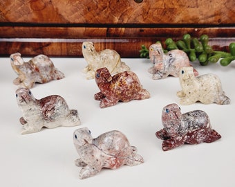 May include: A collection of hand-carved stone seal figurines in various colors, including cream, red, and gray. Each seal has blue eyes and is posed in a resting position. The figurines are displayed on a white surface.