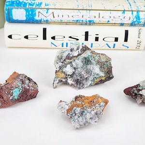 Op de afbeelding: Vier ruwe mineraalmonsters met verschillende kleuren en texturen. De mineralen vertonen tinten bruin, rood, wit en turquoise. De achtergrond bevat twee boeken met de titels "Mineralogy" en "Celestial Minerals."