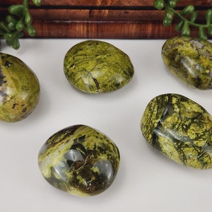 May include: Five polished green serpentine stones with dark veining. The stones are oval-shaped and vary in size. The background is a white surface with a wooden backdrop and greenery.