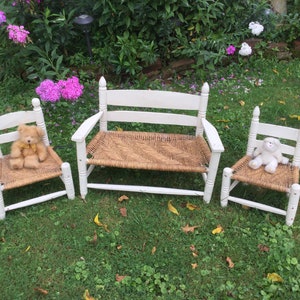 May include: A set of three white wooden chairs with woven rush seats. The chairs are arranged in a row on a grassy lawn. A teddy bear sits on one chair and a white stuffed animal sits on another.