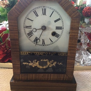 May include: A vintage wooden clock with Roman numeral markings on the face. The clock has a black and gold design on the bottom and a wood grain finish.