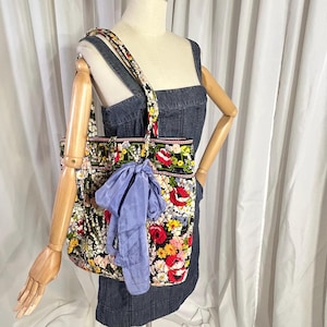 Puede incluir: Un bolso tote con estampado floral con una correa a juego y una bufanda azul claro atada al asa. El bolso presenta un fondo negro con flores rojas, amarillas y blancas. El bolso se muestra sobre un vestido vaquero.