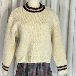 May include: Cream-colored sweater with a ribbed hem and cuffs. The sweater features a brown and pink striped collar and matching stripes on the cuffs. The sleeves are long, and the sweater has a relaxed fit. The fabric appears to be a soft knit.