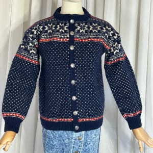 Könnte beinhalten: Eine marineblaue Strickjacke mit einem Schneeflocken- und geometrischen Muster in Weiß und Rot. Der Pullover hat einen runden Kragen, lange Ärmel und silberne Knopfverschlüsse. Die Strickjacke wird auf einer Schaufensterpuppe präsentiert.
