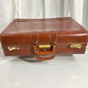 Puede incluir: Una maleta rectangular de color marrón con acabado liso y costuras blancas en contraste. Cuenta con un asa central, dos cierres metálicos de tono dorado en la parte frontal y un pequeño emblema circular debajo del asa.