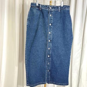 May include: A medium-wash denim skirt with a button-down front. The skirt is knee-length and hangs on a hanger. The skirt has a classic five-pocket design and a button and zipper closure at the waist. The skirt is made of a sturdy denim fabric.