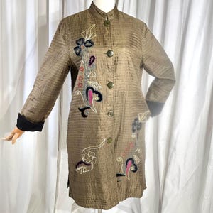May include: A long, olive green coat with a mandarin collar and decorative floral embroidery. The coat features a quilted texture, long sleeves with black cuffs, and green buttons down the front. The embroidery includes black, pink, and white accents.