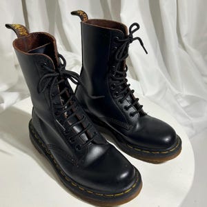 May include: A pair of black leather lace-up boots with a classic design. The boots have a tall shaft, a rounded toe, and a yellow stitched sole. The interior is brown. The boots are on a white surface.
