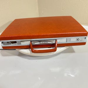May include: A rectangular orange hardshell briefcase with a textured finish. The case features a silver-toned metal frame, a matching orange handle, and two key-operated locking mechanisms. The brand name Samsonite is printed on the handle.