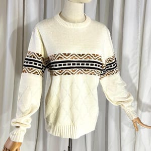 May include: Cream-colored sweater with a crew neck and long sleeves. The sweater features a geometric pattern in brown and black across the chest and sleeves. The lower portion of the sweater has a diamond pattern.