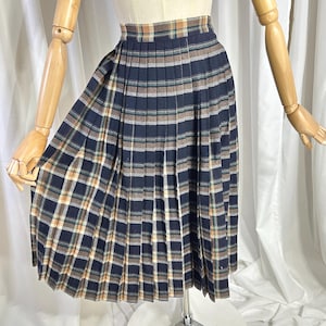 Puede incluir: Una falda plisada hasta la rodilla con un patrón de cuadros en azul marino, beige y verde apagado. La falda tiene una cintura ajustada y un diseño de pliegues uniformes y estructurados que se extienden desde la cintura hasta el dobladillo.