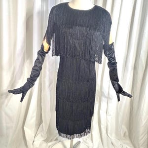 Könnte beinhalten: Ein schwarzes ärmelloses Kleid mit mehreren horizontalen Lagen aus langen Fransen, die das gesamte Kleidungsstück vom Ausschnitt bis zum Saum bedecken. Dazu gehört ein Paar passende, ellenlange Handschuhe aus schwarzem Satin.