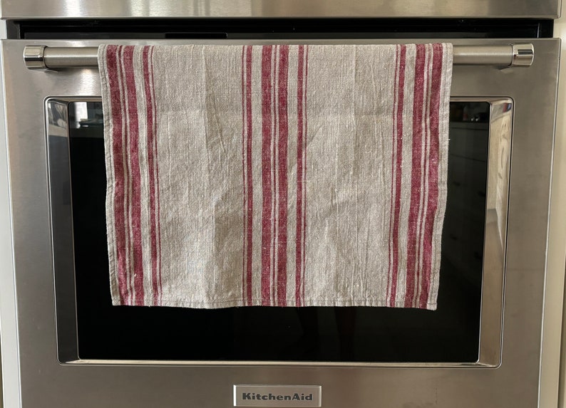 May include: A gray and burgundy striped kitchen towel hanging on a silver oven door handle. The oven door is closed and the brand name "KitchenAid" is visible on the bottom of the door.