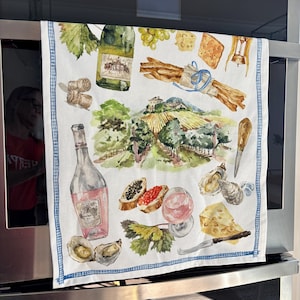 May include: A white kitchen towel with a blue and white checkered border. The towel features a watercolor illustration of a vineyard, wine bottles, grapes, cheese, breadsticks, and oysters.