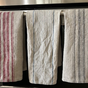 May include: Three striped kitchen towels hanging on a rack. The towels are made of a linen blend and have different colored stripes. The towels are hanging in front of a stainless steel oven.