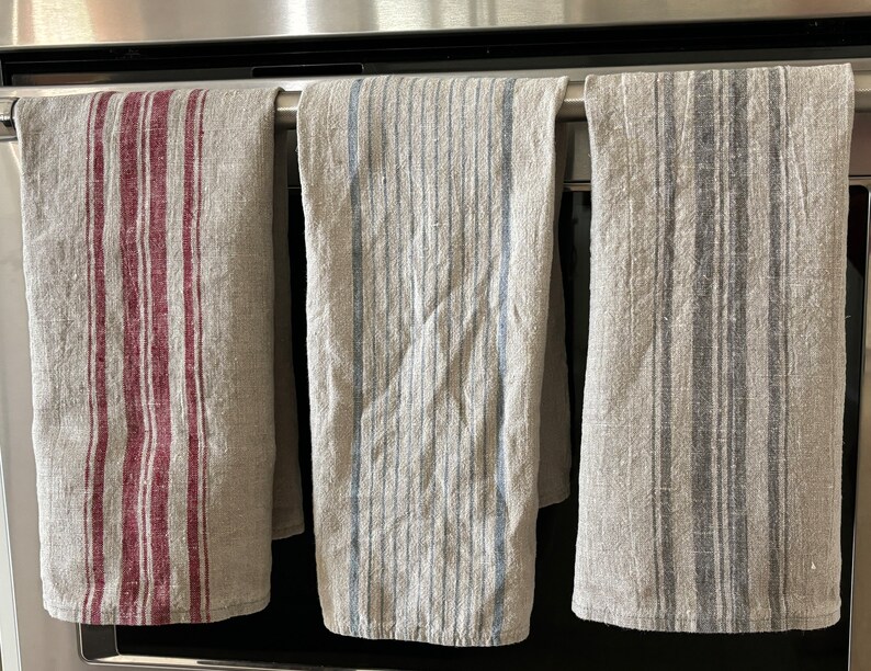 May include: Three striped kitchen towels hanging on a rack. The towels are made of a linen blend and have different colored stripes. The towels are hanging in front of a stainless steel oven.