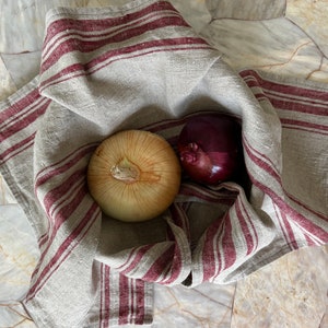 May include: A white onion and a red onion are nestled in a linen kitchen towel with burgundy stripes. The towel is folded over the onions, creating a rustic and cozy feel.