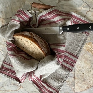 May include: A loaf of bread, sliced in half, is placed inside a linen kitchen towel with red and white stripes. A serrated bread knife with a black handle is lying on top of the towel.