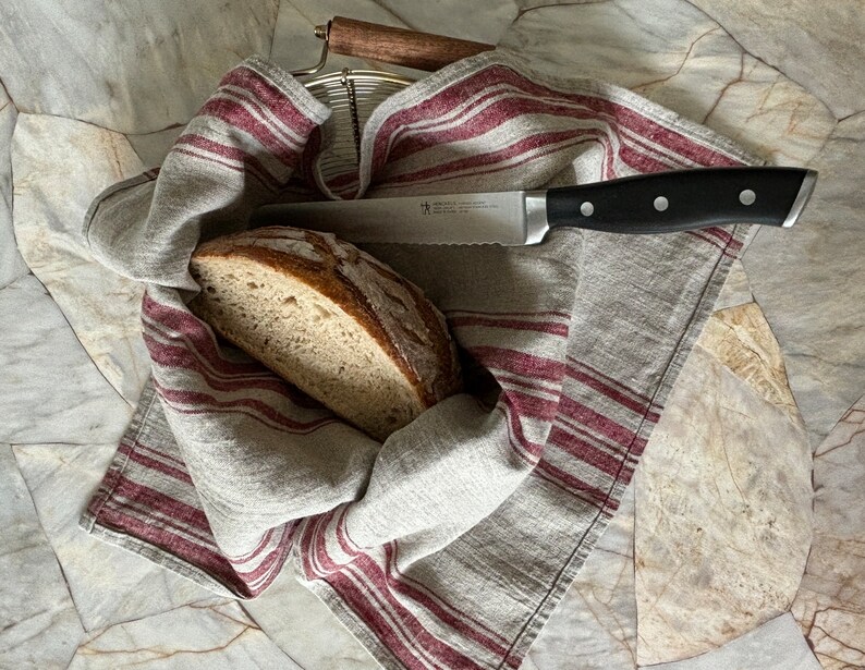 May include: A loaf of bread, sliced in half, is placed inside a linen kitchen towel with red and white stripes. A serrated bread knife with a black handle is lying on top of the towel.