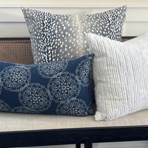 May include: Three decorative cushions in a home setting. One is navy blue with white floral patterns, another is white with a ribbed texture, and the third is grey with a white spotted design. The cushions are arranged on a neutral-toned bench.