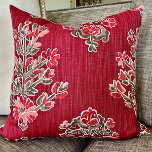 Pode incluir: Uma almofada decorativa quadrada com um fundo vermelho vibrante e um padrão floral em tons de vermelho, rosa e cinza. A almofada é colocada em um sofá cinza, mostrando seu design e textura. As dimensões da almofada não são visíveis.