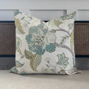 May include: A square decorative pillow with a floral pattern in shades of blue, green, and beige. The pillow features large, stylized flowers and leaves on a light background. The pillow is placed on a neutral-colored surface.