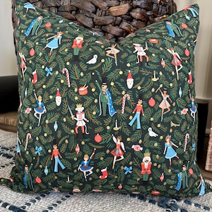 Puede incluir: Un cojín cuadrado decorativo con un fondo verde oscuro que presenta un patrón repetido de ilustraciones navideñas. El diseño incluye cascanueces, bailarinas, bastones de caramelo y adornos. El cojín está colocado sobre una superficie tejida.