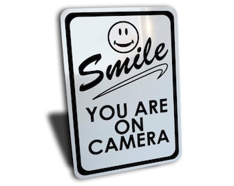May include: A silver metal sign with black text and a smiley face. The sign reads "Smile You Are On Camera".