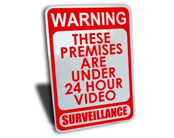 Op de afbeelding: Een rood en wit waarschuwingsbord met de tekst "WARNING THESE PREMISES ARE UNDER 24 HOUR VIDEO SURVEILLANCE".