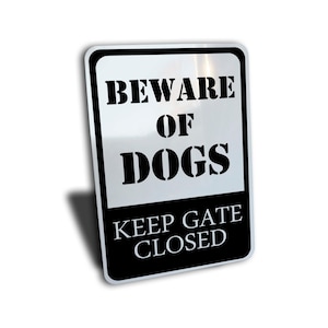 Op de afbeelding: Een zwart-wit metalen bord met de tekst "Beware of Dogs Keep Gate Closed".