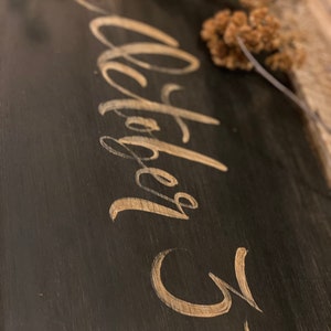 May include: A dark wooden sign with the words "October 31" in elegant gold script. The sign has a rustic, distressed appearance, with visible wood grain. Dried flowers are arranged at the top, adding a natural touch to the design.