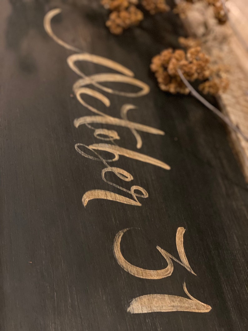 May include: A dark wooden sign with the words "October 31" in elegant gold script. The sign has a rustic, distressed appearance, with visible wood grain. Dried flowers are arranged at the top, adding a natural touch to the design.