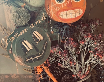 May include: Two vintage Halloween decorations, one orange and one green, with painted faces and large eyes. The decorations are in a brown and white ceramic vase with dried flowers and pine cones.