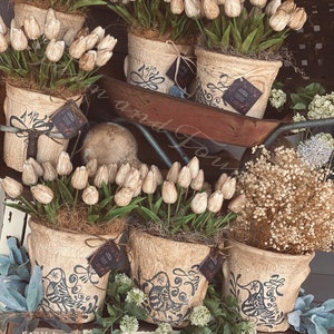 May include: An arrangement of potted faux tulips in rustic, textured pots. The pots are adorned with dark blue decorative patterns and tags. The tulips are cream-colored with green stems, creating a farmhouse aesthetic.