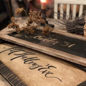 May include: Two rustic wooden signs with black lettering. The top sign reads "October 31" and the bottom sign says "All Hallow's Eve". The signs have a distressed, weathered look with a dark brown frame. Dried flowers and a candle are in the background.
