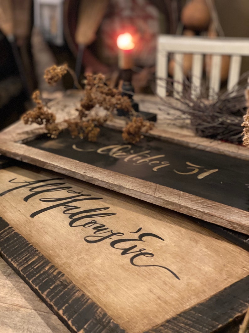 May include: Two rustic wooden signs with black lettering. The top sign reads "October 31" and the bottom sign says "All Hallow's Eve". The signs have a distressed, weathered look with a dark brown frame. Dried flowers and a candle are in the background.