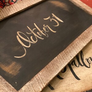 May include: Two rustic wooden framed chalkboards. The top chalkboard is black with the words "October 31" in gold script. The frame is a light brown with a distressed finish. The bottom chalkboard is tan with black script.