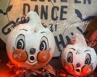 May include: Two vintage-style Halloween decorations. The white, gourd-shaped figures have black eyes, orange cheeks, and open mouths. They are adorned with orange and black paper fringe and have wire handles. The background features the words "HALLOWEEN DANCE".