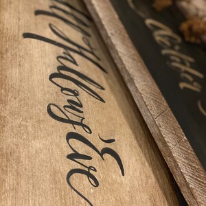 May include: Two wooden signs with black calligraphy text. The first sign has the words "Happy Life" written in a flowing script. The second sign has the word "Wild" written in a similar style. The signs have a rustic, farmhouse aesthetic.