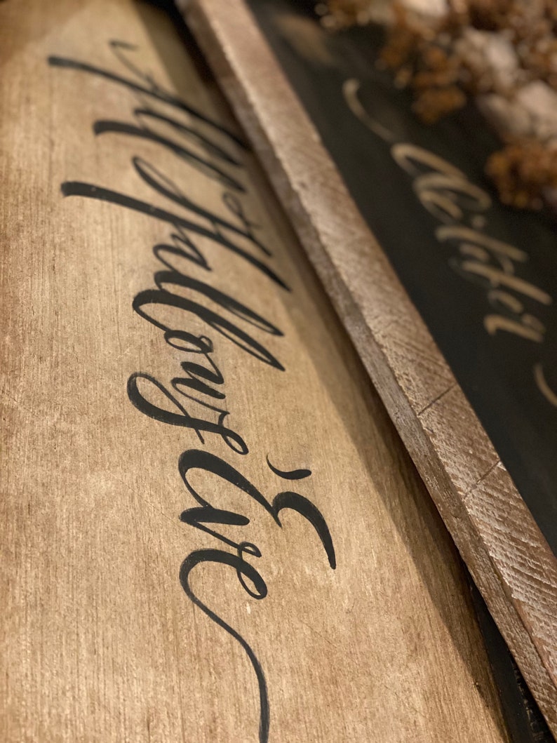 May include: Two wooden signs with black calligraphy text. The first sign has the words "Happy Life" written in a flowing script. The second sign has the word "Wild" written in a similar style. The signs have a rustic, farmhouse aesthetic.